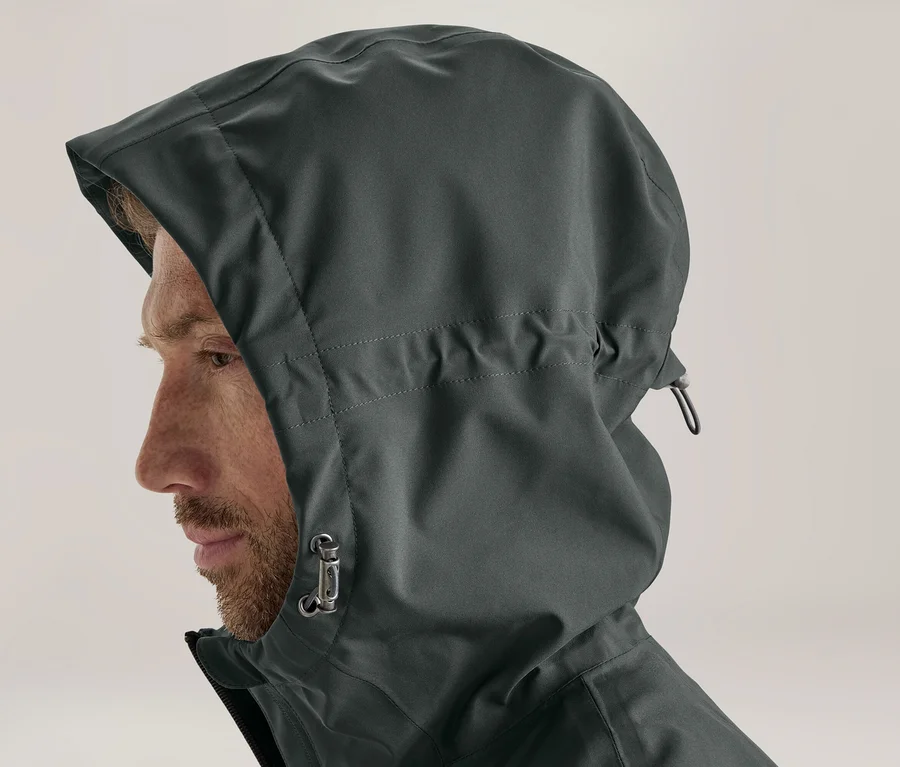 Mann im Profil trägt eine dunkelgraue Kapuzenjacke mit hochgezogener Kapuze. Sein bärtiges Gesicht ist sichtbar, die Kapuze hat einen Kordelzug und Stopper.