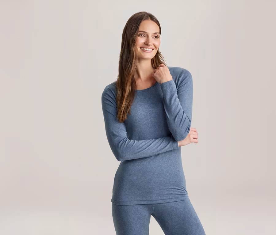 Eine lächelnde Frau mit langen braunen Haaren trägt ein blaues Langarmshirt und passende Leggings und blickt nach rechts.