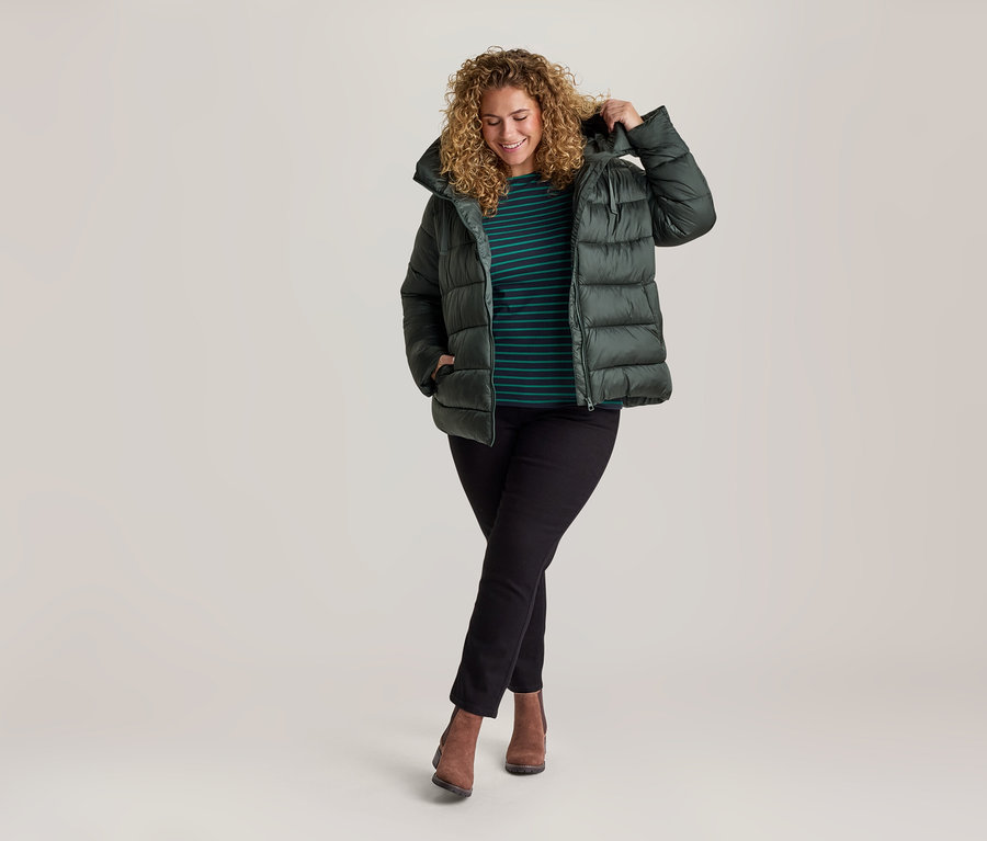 Lächelnde Frau mit lockigem blondem Haar, trägt eine dunkelgrüne Steppjacke, gestreiftes Oberteil, schwarze Straight Jeans – Fit »Juna« und braune Stiefeletten, steht mit gekreuzten Beinen.