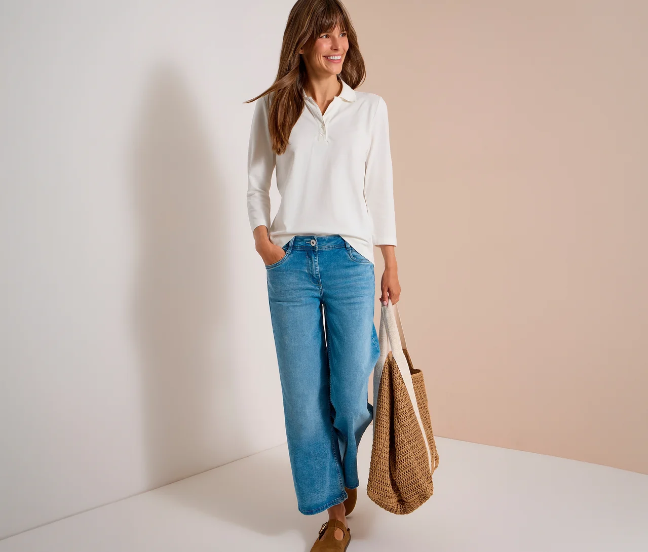 Lächelnde Frau in weißem CECIL 3/4-Poloshirt und blauen Jeans, eine Strohtasche tragend.