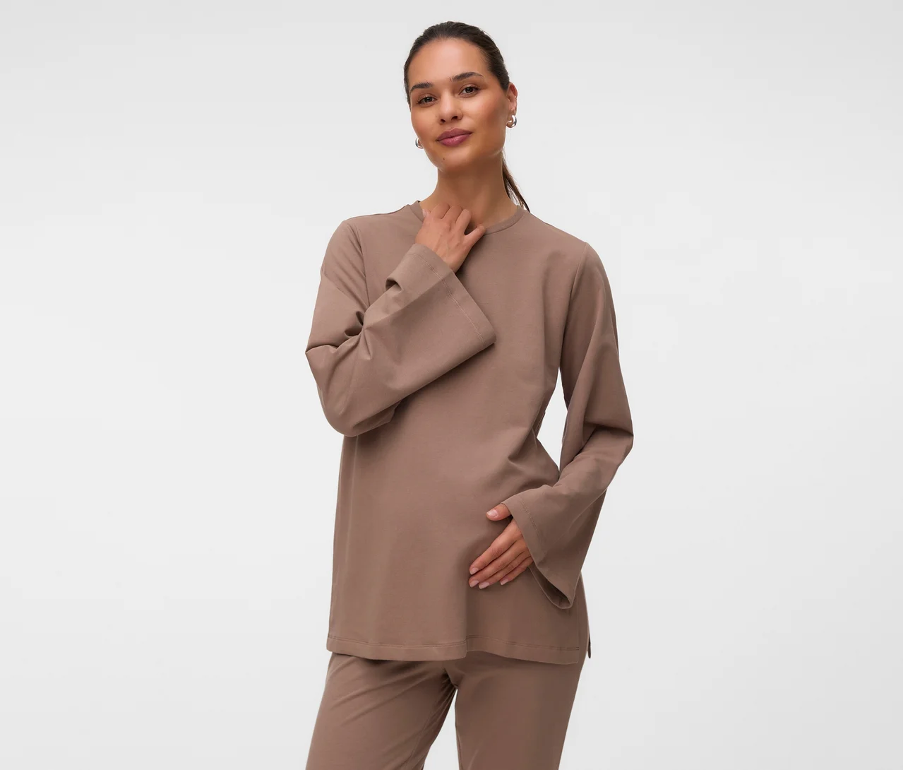 Eine Frau trägt ein hellbraunes MAMALICIOUS Sweat Top »MLSINE LS« und passende Hose, ihre linke Hand liegt auf ihrem schwangeren Bauch, die rechte am Hals, während sie in die Kamera blickt.