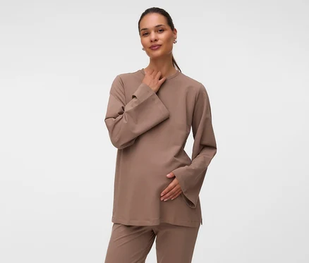 Eine Frau trägt ein hellbraunes MAMALICIOUS Sweat Top »MLSINE LS« und passende Hose, ihre linke Hand liegt auf ihrem schwangeren Bauch, die rechte am Hals, während sie in die Kamera blickt.