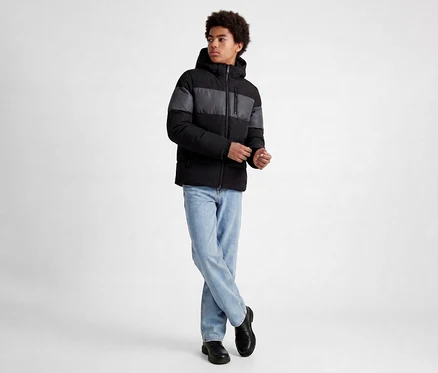 Junger Mann mit lockigem Haar trägt eine schwarz-grau gestreifte Steppjacke, hellblaue Jeans und schwarze Schuhe, steht mit gekreuzten Beinen und verschränkten Händen.