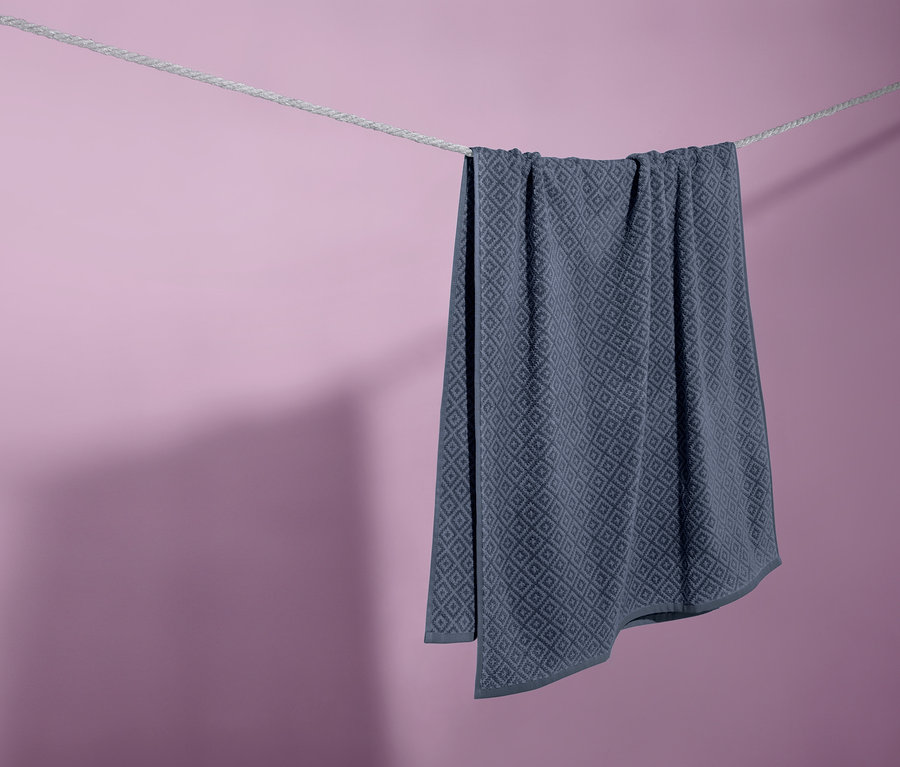 Ein blaues Jacquard-Badetuch hängt an einer Wäscheleine vor einem lila Hintergrund.