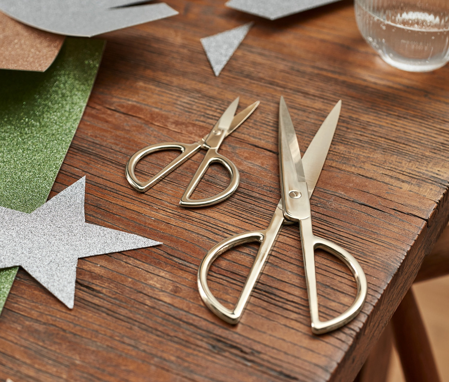 Auf einem Holztisch liegen ein Bastelscheren-Set, bunte Glitzerpapiere und ein Glas Wasser.