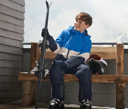 Ein Junge sitzt auf einer Bank in einer Skijacke und hält Ski.