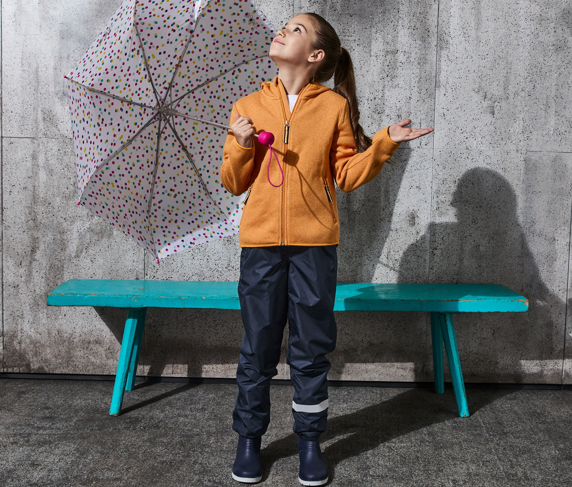 Ein Mädchen schaut nach oben, es steht mit einem Regenschirm und einer Regenhose, packbar, aus recyceltem Material.