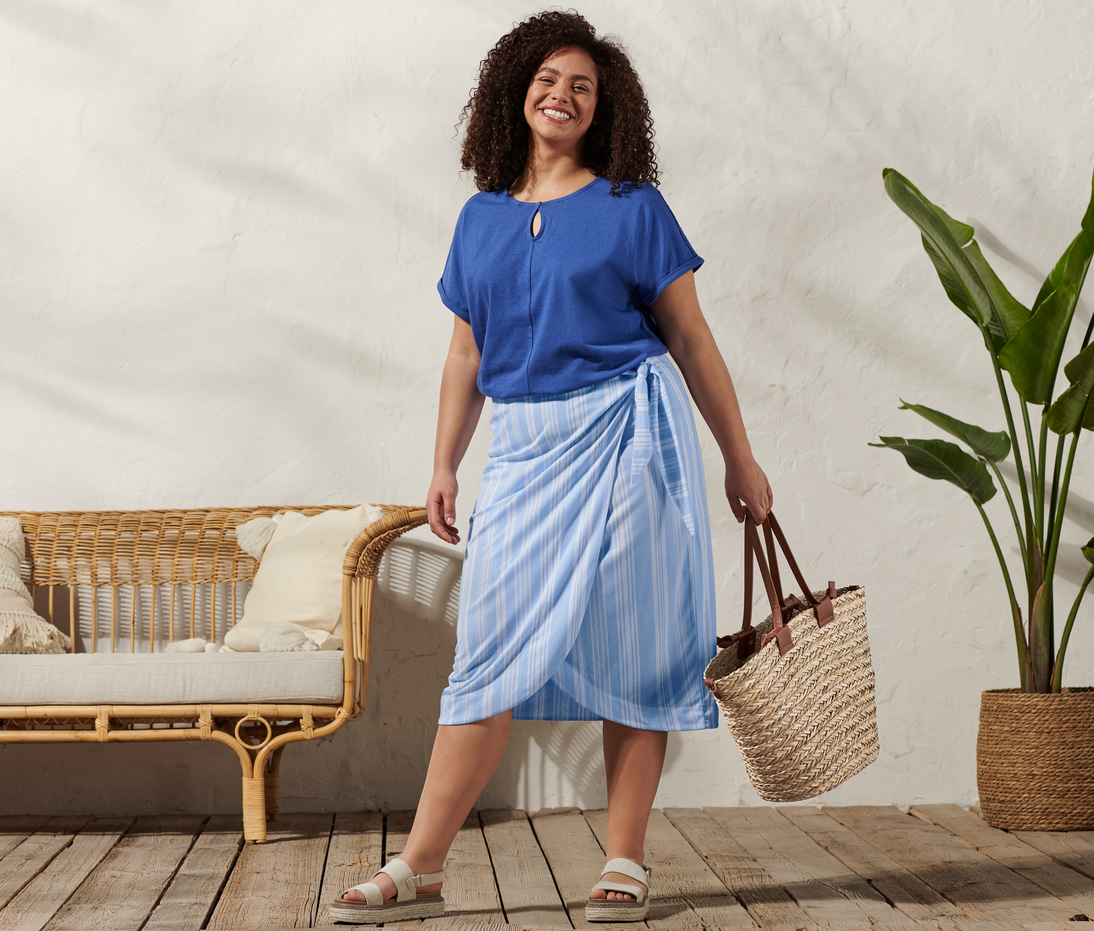 Frau trägt einen Shirt aus Leinenmix, einen gestreiften Wickelrock, Plateau-Sandale aus Leder und eine Korbtasche.