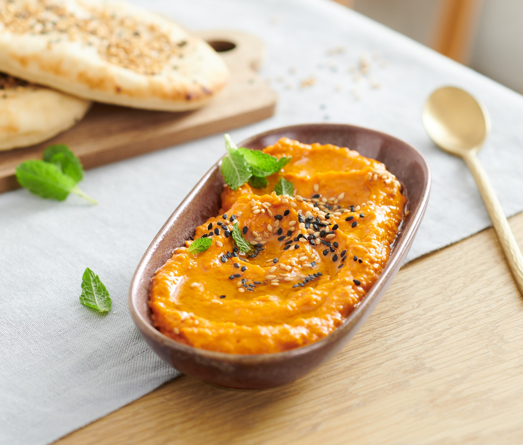 Zwei Brote auf einem Brett neben einer kleinen Servierplatte mit Aufstrich, einem Glas mit Getränk und einem Löffel auf dem Tisch.
