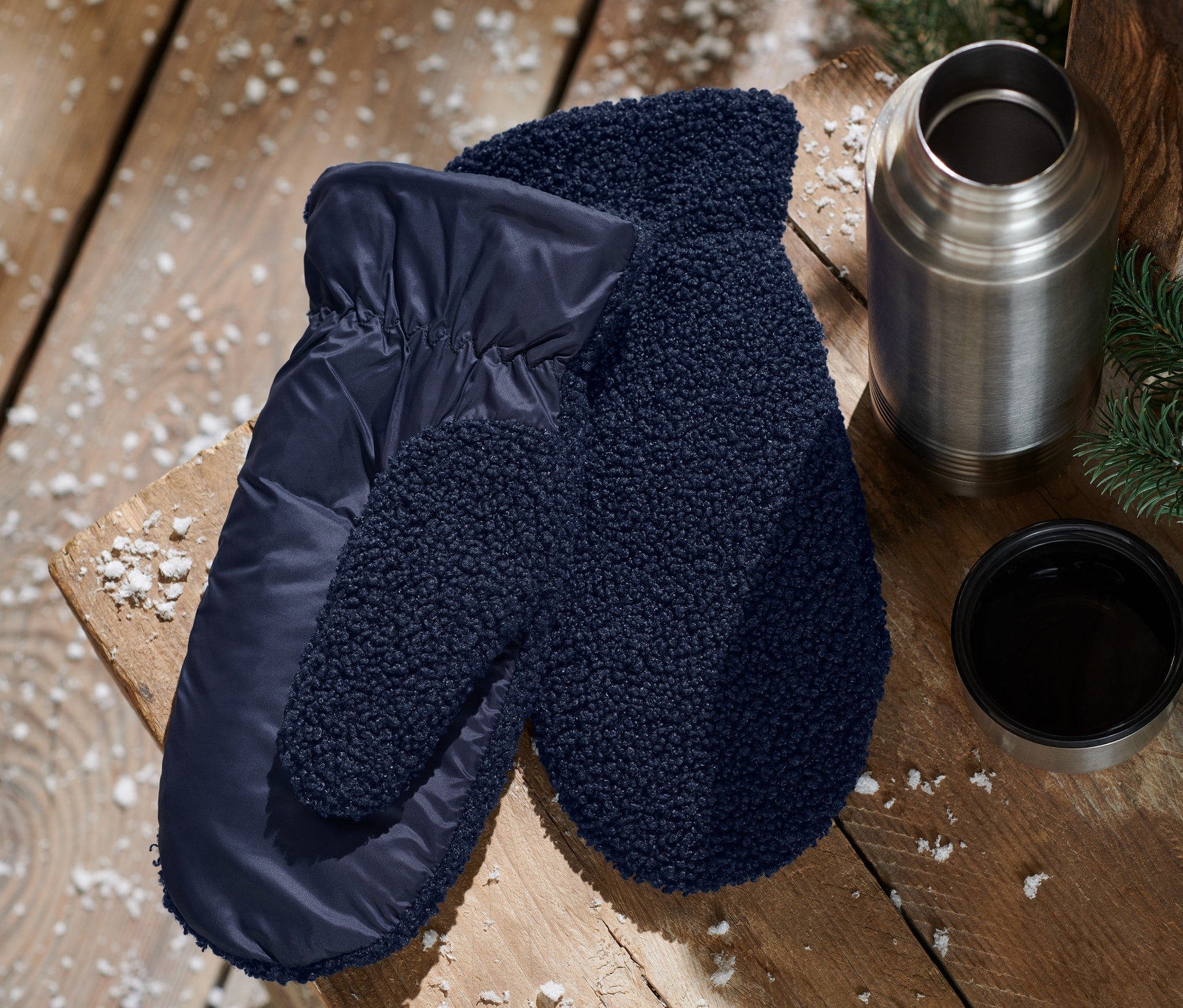 Auf einem Holztisch liegen blaue Fäustlinge, eine Thermoskanne und eine Tasse.