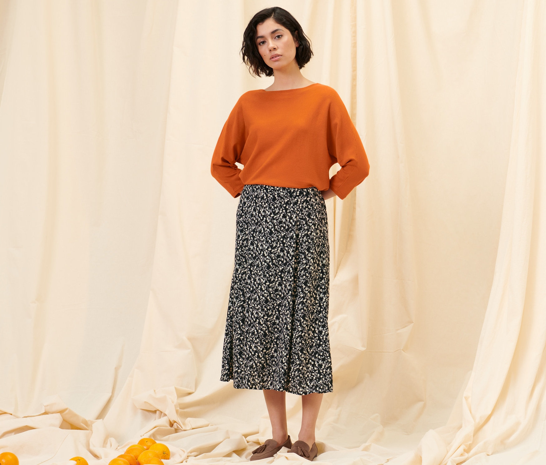 Frau steht und trägt einen floralen Midi-Rock, einen orangefarbenen Feinstrickpullover und Loafer aus Veloursleder.
