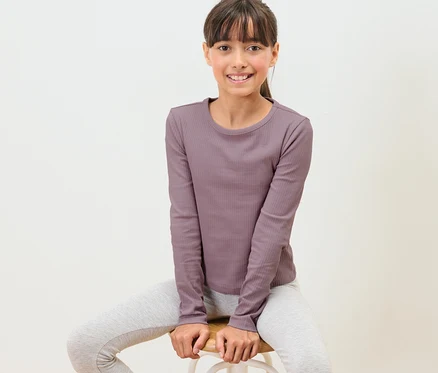 Mädchen sitzt auf einem Hocker und trägt ein Kinder-Langarmshirt mit Rippstruktur.