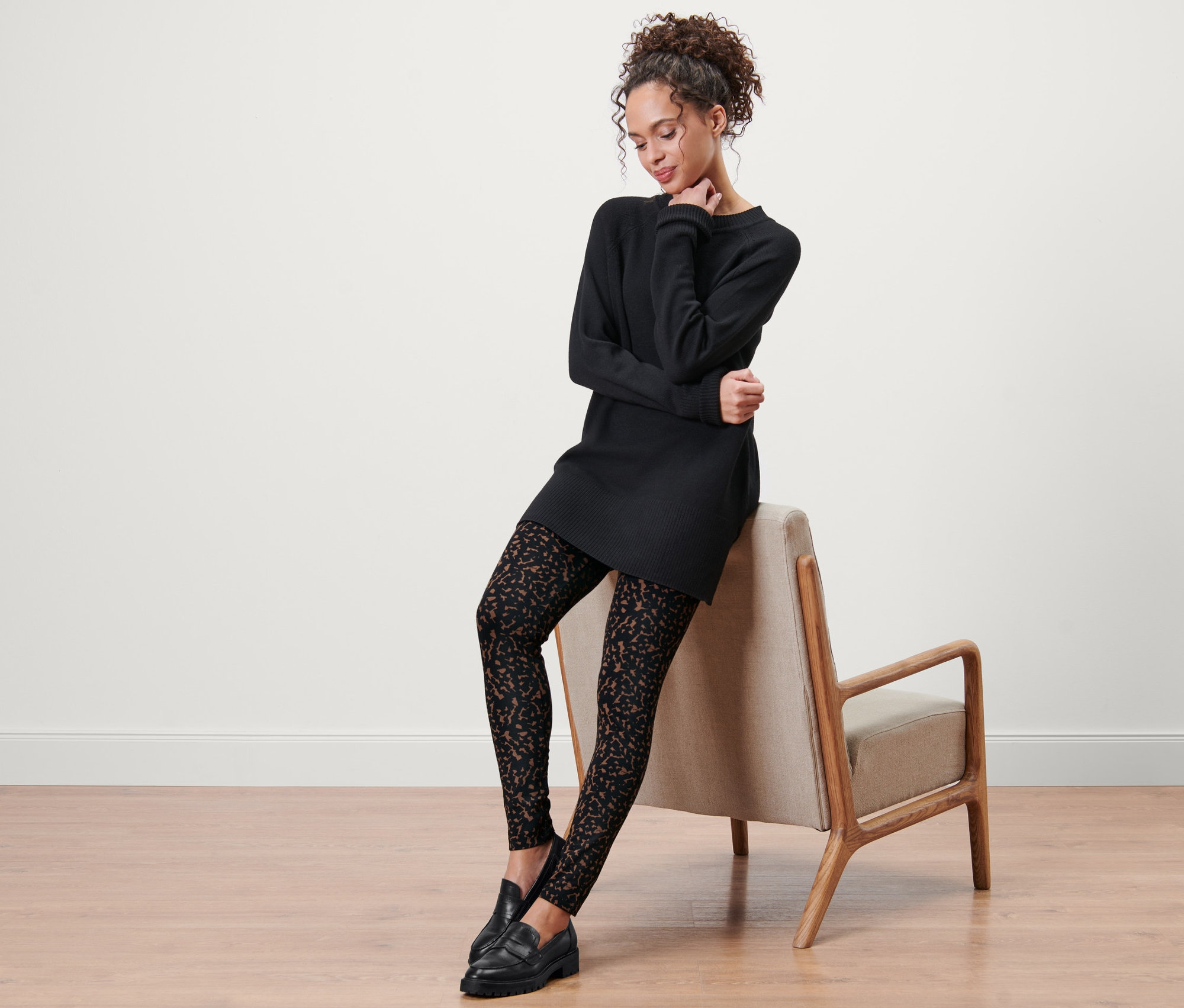 Eine Frau sitzt auf einem Stuhl und trägt ein schwarzes Kleid und bedruckte Leggings.