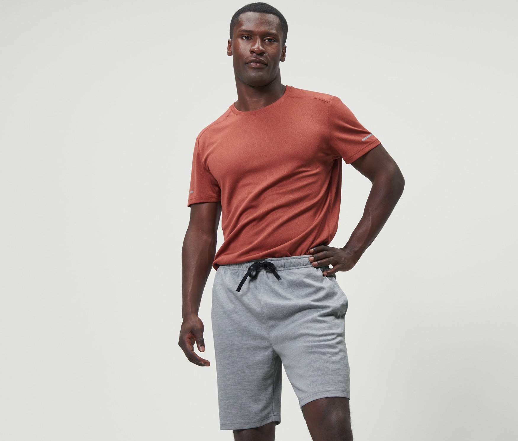 Mann posiert in grauen Sportshorts und orangefarbenem T-Shirt.