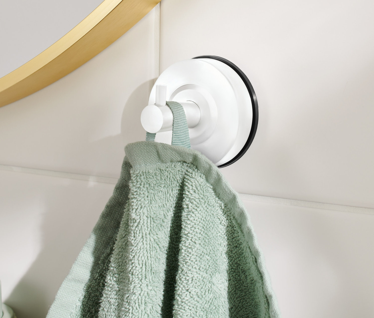 An einer weißen Wand hängt ein Haken mit Saugnapf, an dem ein grünes Handtuch hängt. Im Hintergrund sind ein goldener Spiegelrahmen und ein grüner Seifenspender zu sehen.