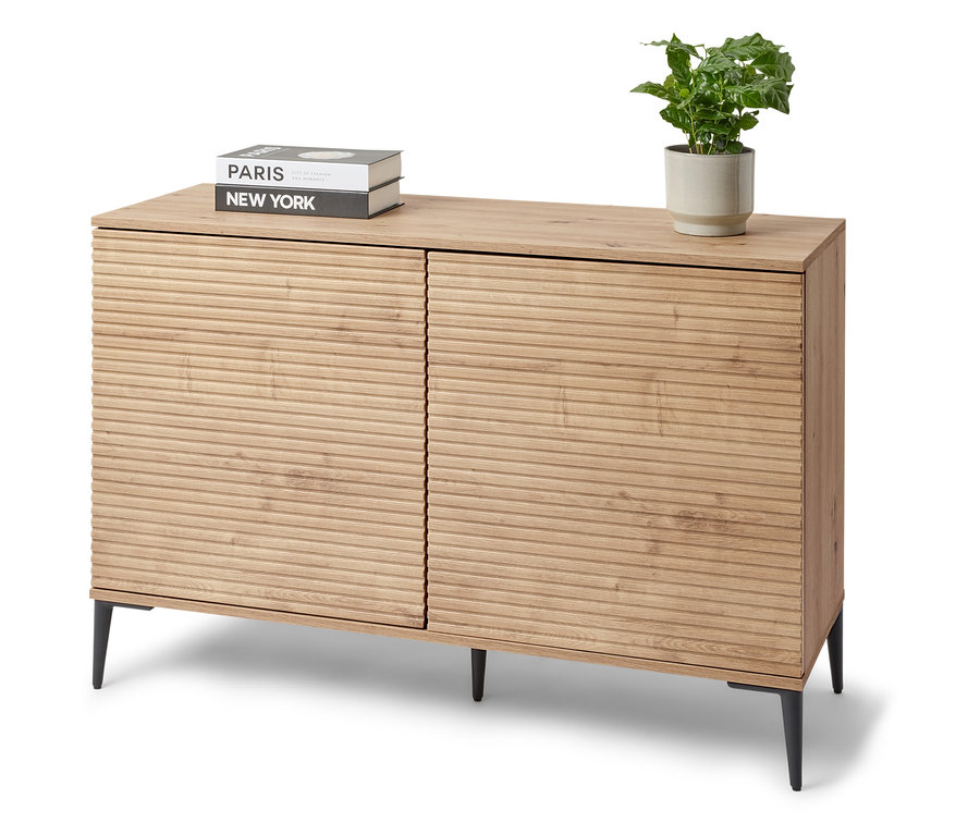 Ein Sideboard »REET« mit Lamellenoptik steht im Raum. Auf dem Sideboard liegen Bücher und eine Topfpflanze.