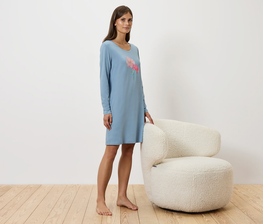 Frau steht neben einem weißen Sessel und trägt ein blaues Nachthemd.