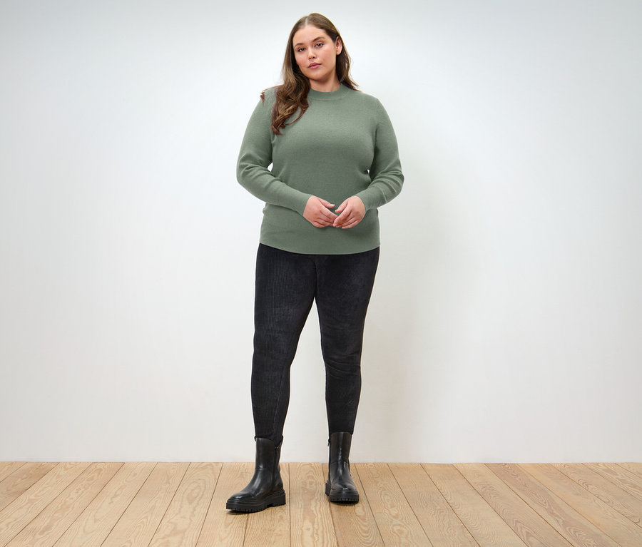 Eine Frau steht auf einem Holzboden und trägt einen grünen Pullover, schwarze Jeans und schwarze Stiefel.