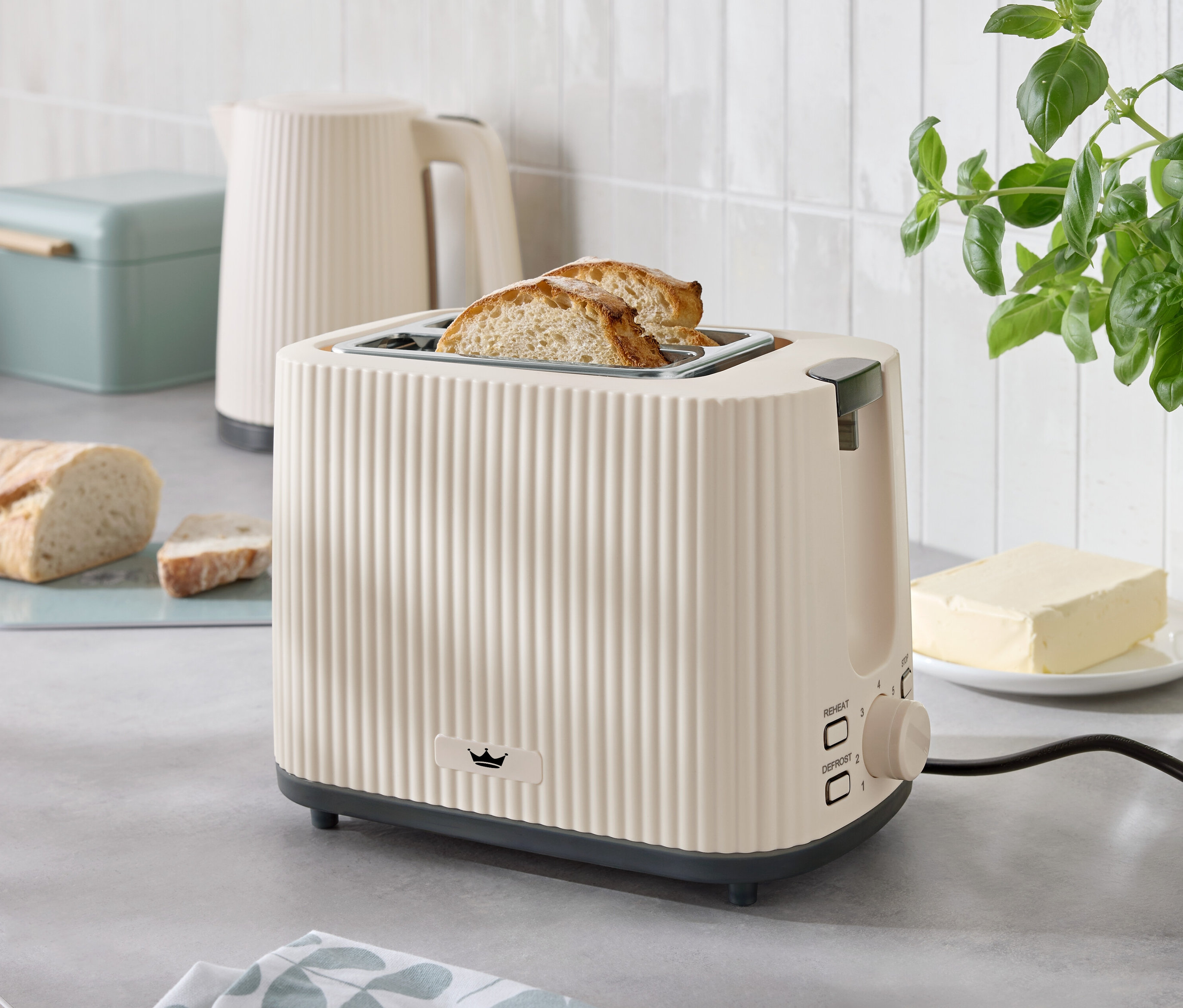 Zwei Toastscheiben ragen aus einem beigen Toaster auf einer Küchentheke, neben einem Wasserkocher, einer Butterdose und einem Brotkasten.