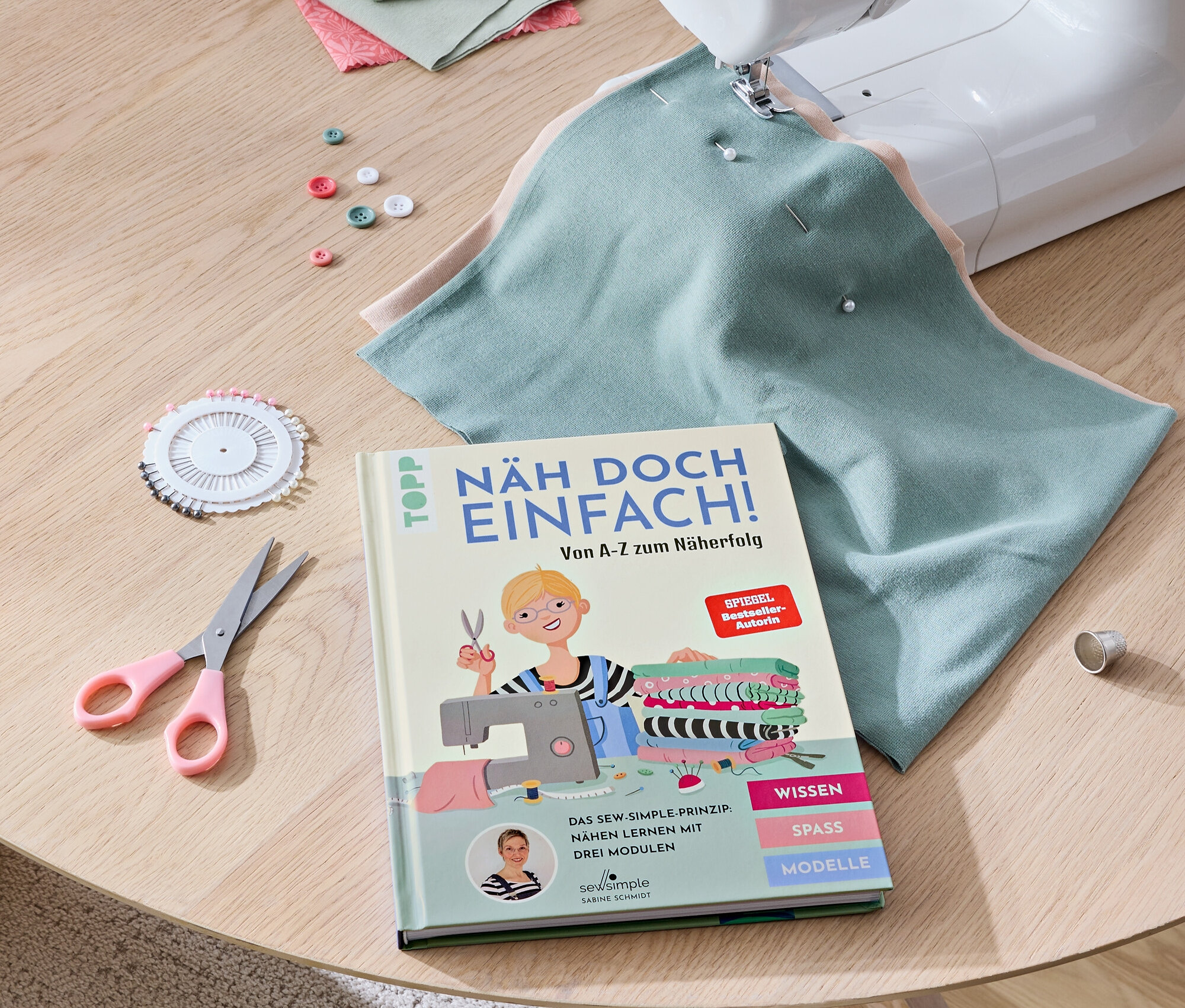 Auf einem Tisch liegen eine Nähmaschine, das Buch »Näh doch einfach«, Stoffe, eine Schere, Knöpfe und Stecknadeln.