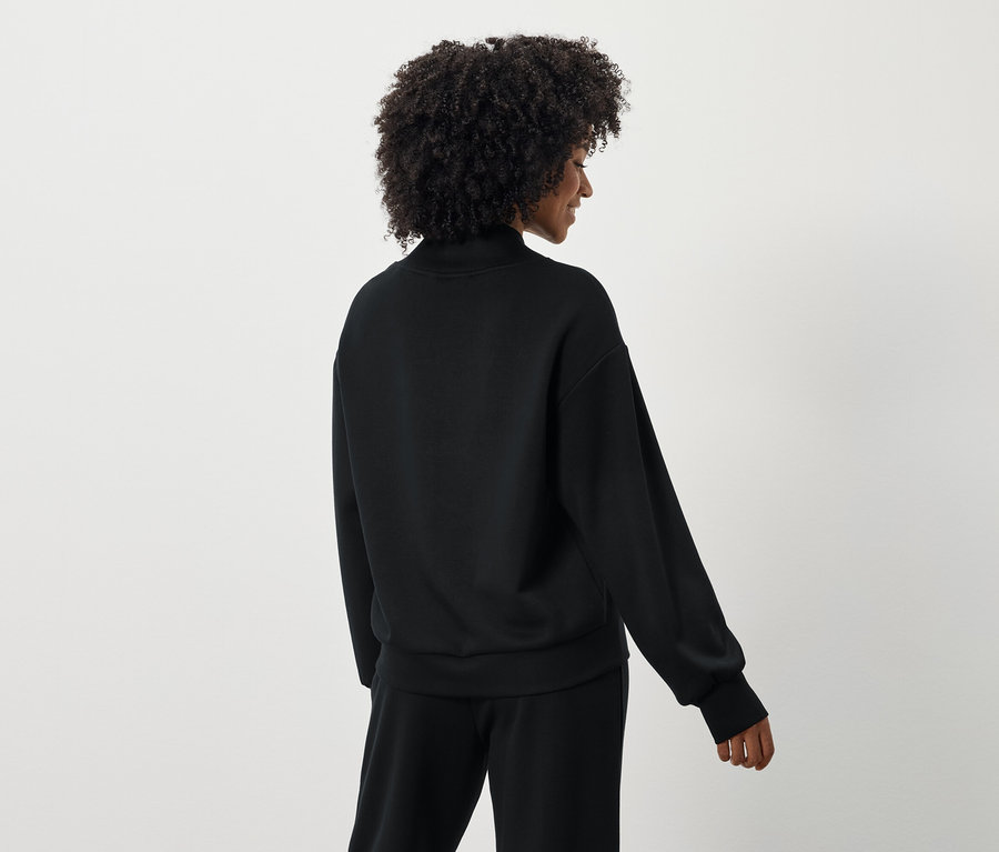 Rückansicht einer Frau mit lockigem Haar, die einen schwarzen Pullover und eine Hose trägt.