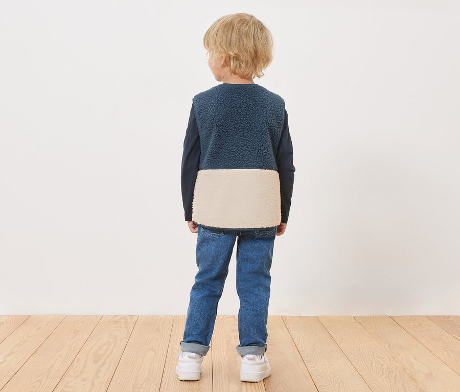 Junge steht mit dem Rücken zur Kamera und trägt eine blaue Weste mit cremefarbenem Unterteil, blaue Jeans und weiße Turnschuhe.