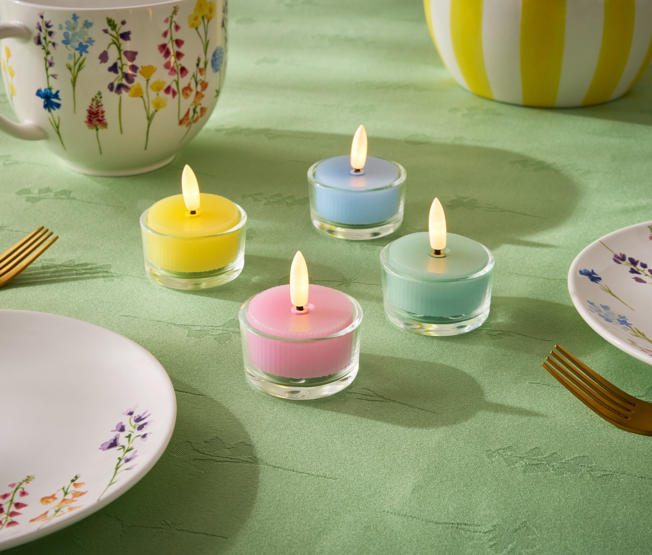 Auf einer grünen Tischdecke stehen vier kleine Kerzen in den Pastellfarben Gelb, Blau, Grün und Rosa. Dazu kommen ein Teller, eine Tasse und eine Schüssel mit Blumenmuster sowie zwei goldene Gabeln.