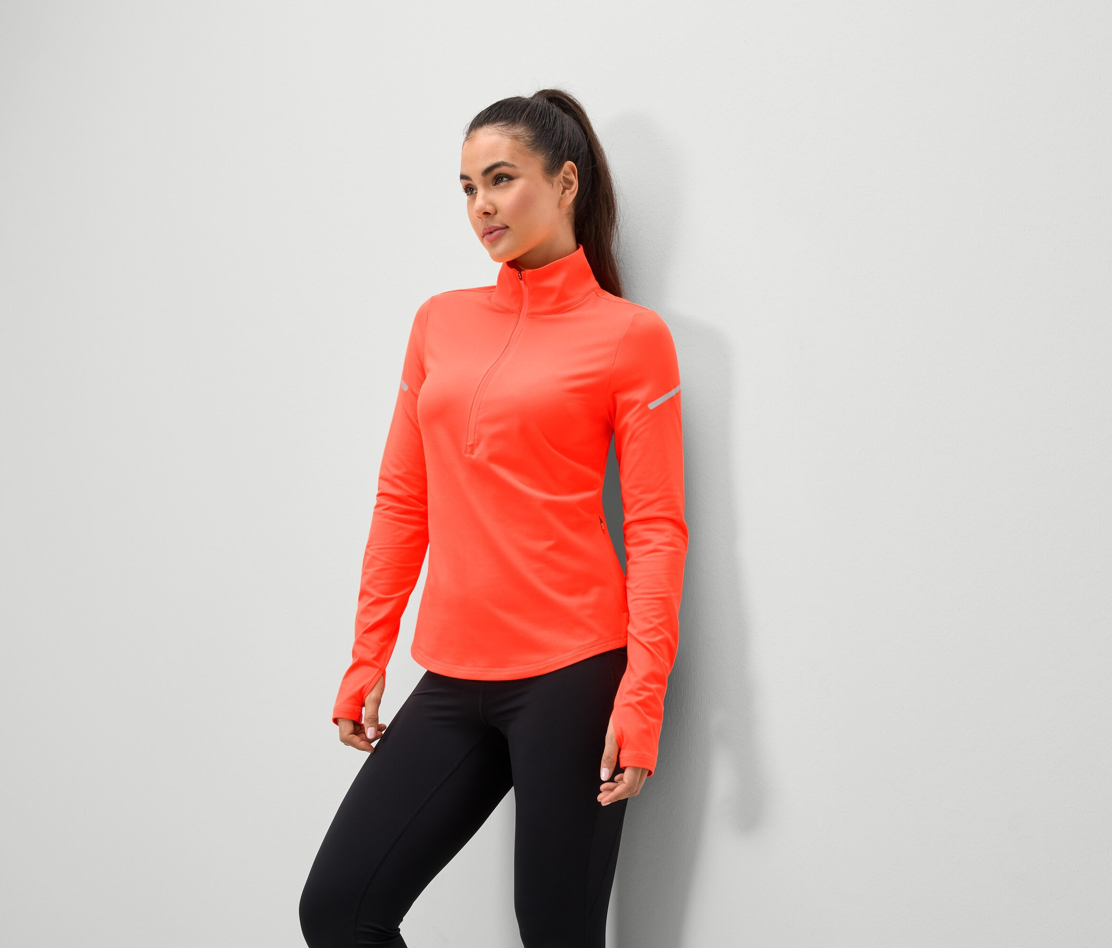 Eine Frau in einer korallenfarbenen Sportjacke mit schwarzen Leggings steht an einer Wand.