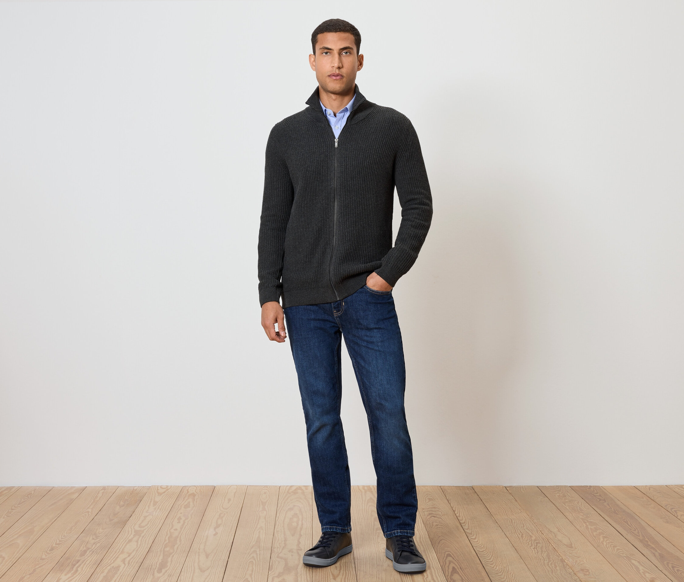 Mann steht und trägt eine Strickjacke, anthrazit meliert, dazu Jeans und Schuhe.