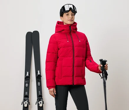 Frau in roter Skijacke hält Skistöcke, daneben Skier und eine Skibrille auf dem Kopf.