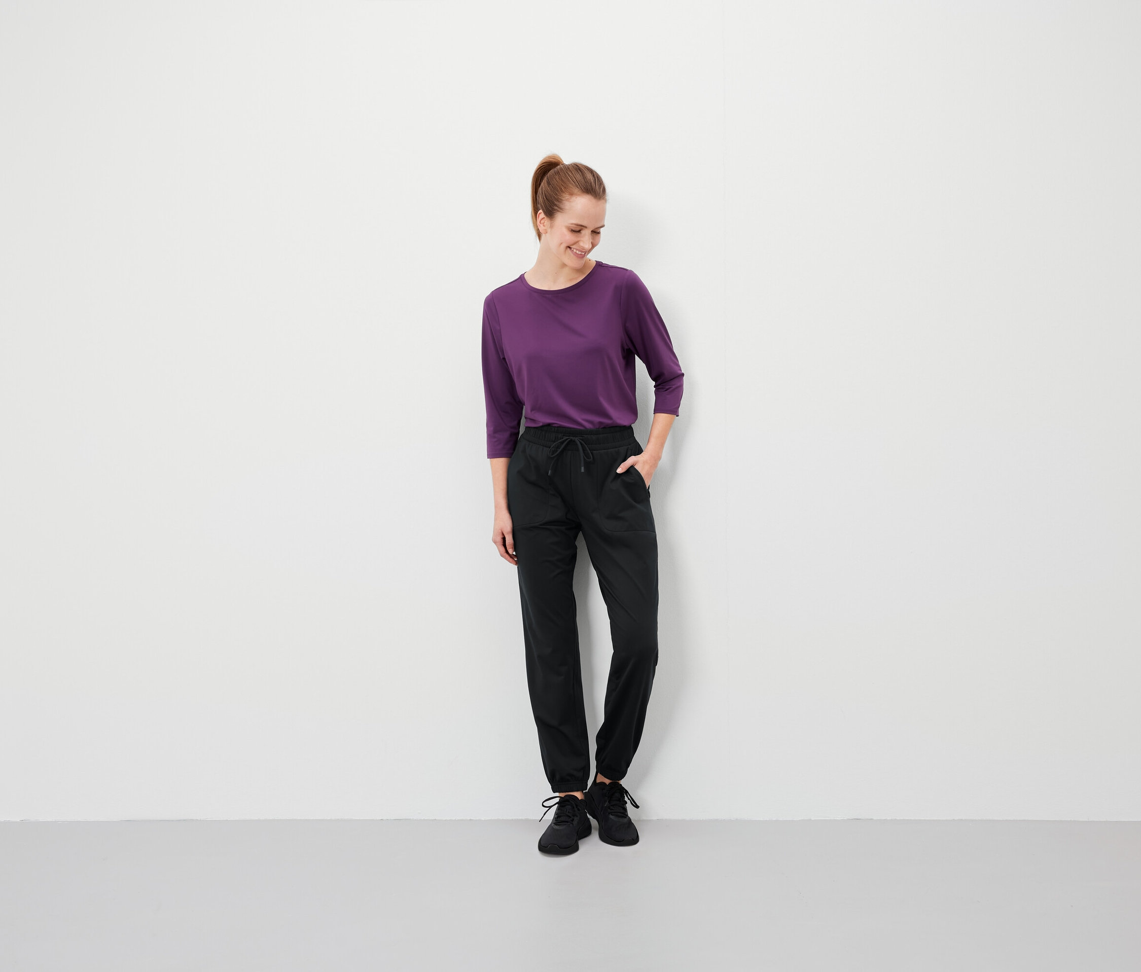 Frau mit braunen Haaren steht mit den Händen in den Taschen ihrer schwarzen Hose und trägt ein violettes T-Shirt.