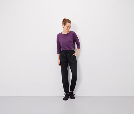 Frau mit braunen Haaren steht mit den Händen in den Taschen ihrer schwarzen Hose und trägt ein violettes T-Shirt.