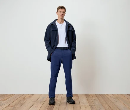 Ein Mann steht auf einem Holzboden. Er trägt eine dunkelblaue Jacke, ein weißes T-Shirt, blaue Hosen und schwarze Schuhe.