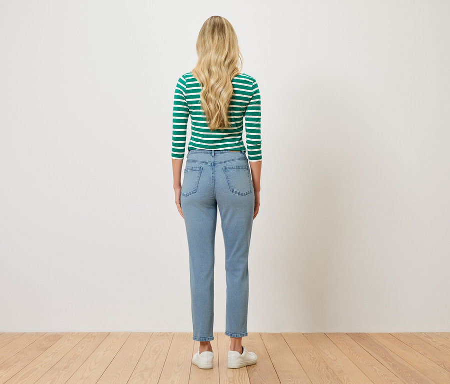 Rückansicht einer Frau mit blonden, gewellten Haaren, die ein grün-weiß gestreiftes Oberteil, hellblaue Jeans und weiße Turnschuhe trägt.