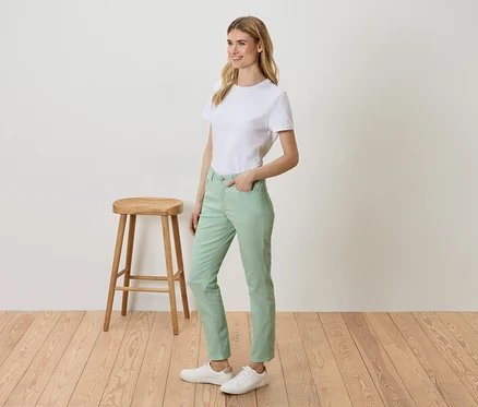 Frau steht neben einem Hocker und trägt ein weißes T-Shirt, grüne Hosen und weiße Turnschuhe.
