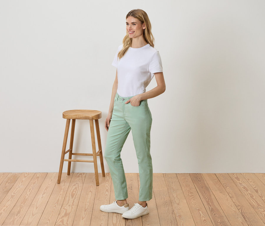 Frau steht neben einem Hocker und trägt ein weißes T-Shirt, grüne Hosen und weiße Turnschuhe.