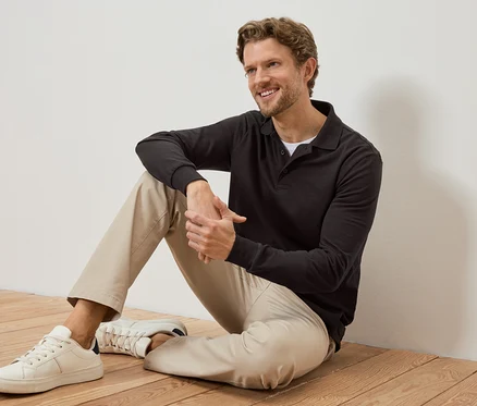 Lächelnder Mann sitzt auf einem Holzboden, trägt ein dunkles Polohemd, helle Hose und weiße Turnschuhe.