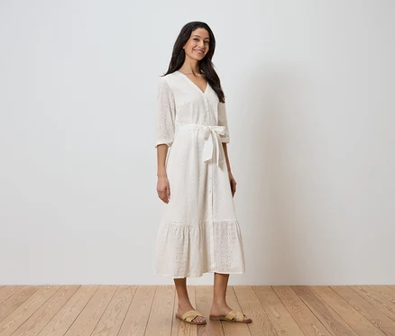 Eine Frau steht vor einer weißen Wand und trägt ein weißes Kleid mit Gürtel und Sandalen.