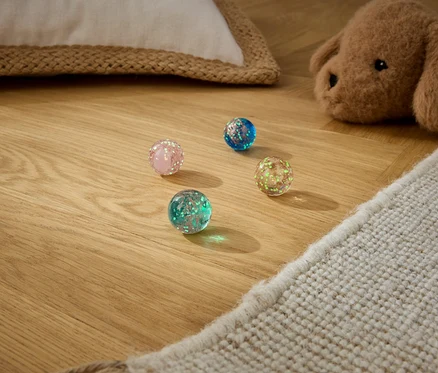 Vier glitzernde Hüpfbälle in Pink, Blau, Grün und Gelbgrün liegen auf einem Holzboden, daneben ein beiger Teppich und ein brauner Stoffhund.