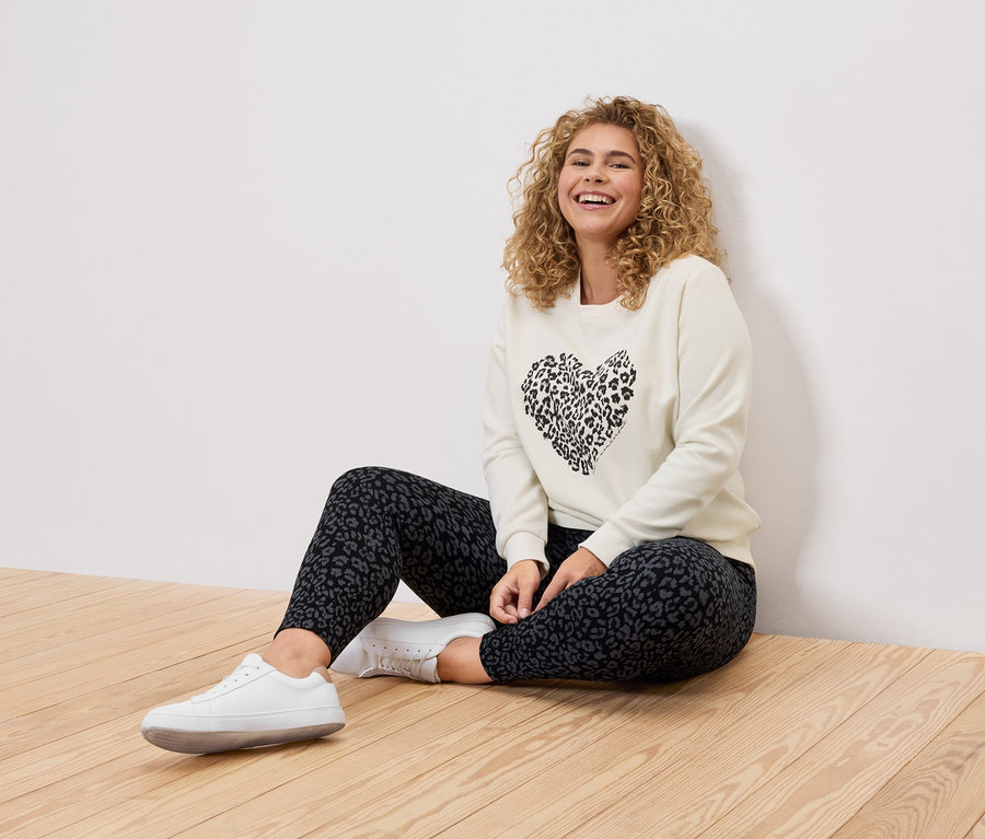 Frau mit lockigem Haar sitzt auf einem Holzboden, gekleidet in einem cremefarbenen Pullover mit einem schwarzen Leopardenherz-Aufdruck, Leggings mit Leopardenmuster und weißen Turnschuhen.