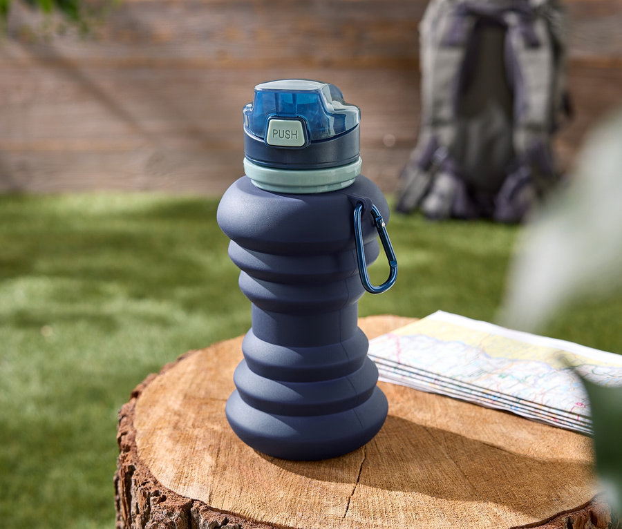 Eine blaue, zusammenfaltbare Wasserflasche mit blauem Verschluss steht auf einem Baumstumpf. Eine Wanderkarte und ein Rucksack sind im Hintergrund zu sehen.