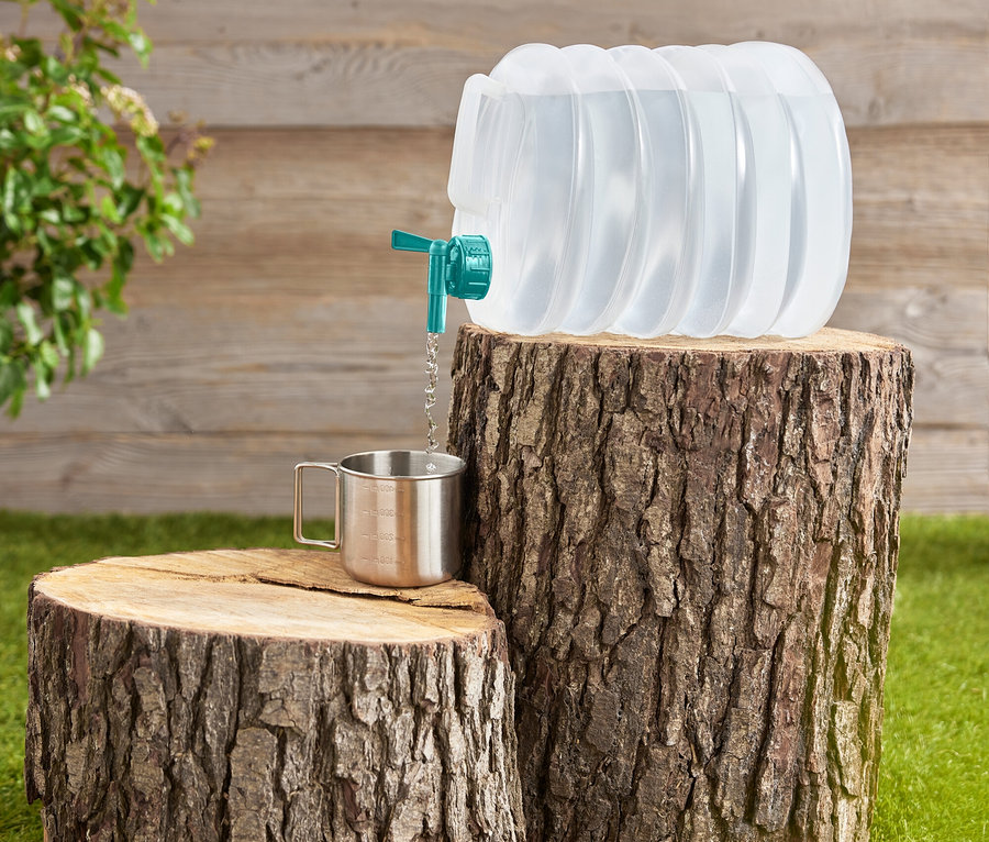 Wasser wird aus einem faltbaren, transparenten Wasserbehälter mit türkisfarbenem Hahn in einen Edelstahlbecher gegossen, der auf Baumstümpfen steht.