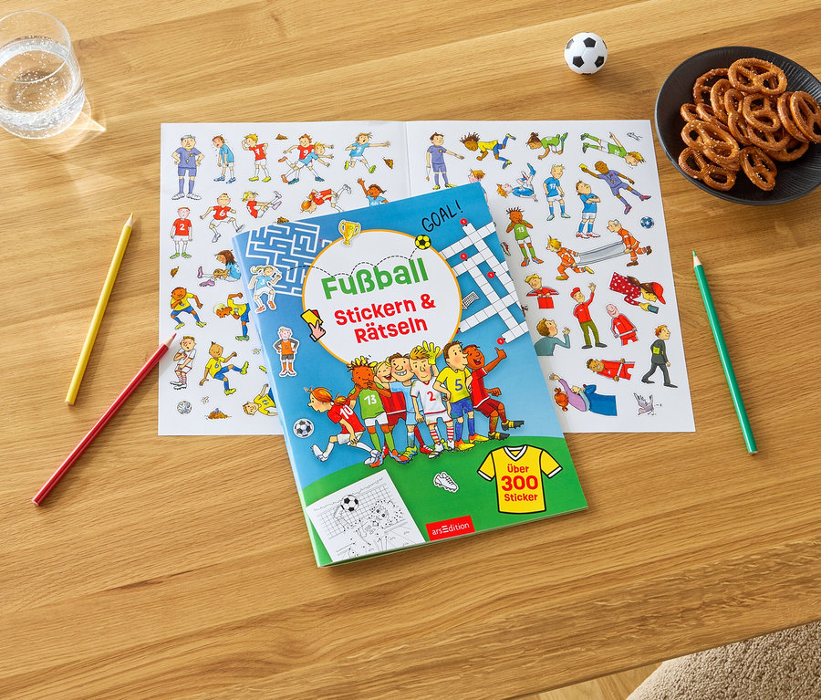 Auf einem Holztisch liegen ein Glas Wasser, eine Schale Brezeln, ein Fußball, Buntstifte und ein Fußball-Sticker- und Rätselbuch mit einem Stickerbogen.