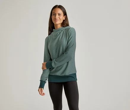 Frau mit langen Haaren trägt einen grün gestreiften Pullover und schwarze Leggings vor einem neutralen Hintergrund.