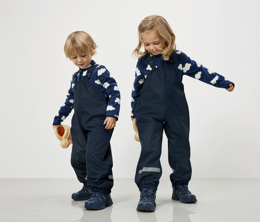 Zwei Kinder in marineblauen Overalls und blau-weißen Wolkenpullovern stehen auf einem weißen Hintergrund.