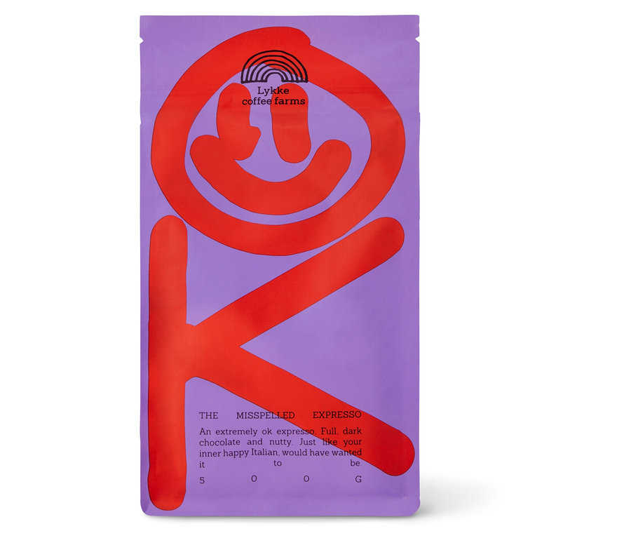 Lila Kaffeepackung mit großem, rotem, stilisiertem „OK“-Grafik. Die Packung zeigt „Lykke coffee farms“ mit einem Regenbogen-Logo, „THE MISSPELLED EXPRESSO“ und eine Beschreibung des Kaffees als „voll, dunkel schokoladig und nussig“. Das Gewicht „500 G“ ist ebenfalls sichtbar.