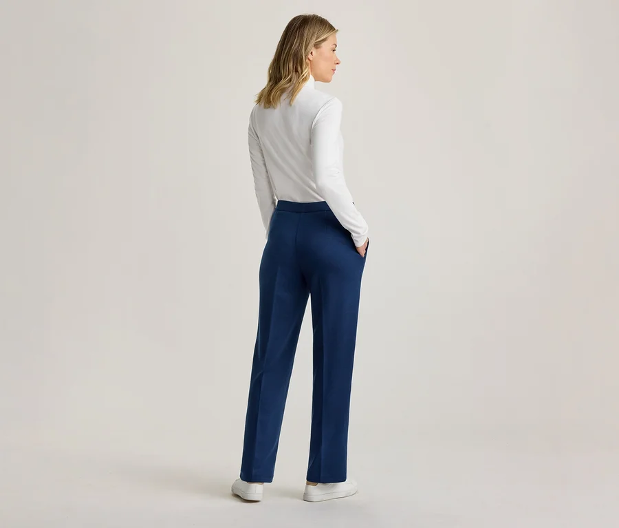 Frau von hinten stehend, trägt ein weißes Langarmshirt und eine blaue Hose, die Hände in den Taschen, blickt nach rechts.