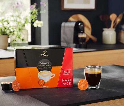 Eine Packung Cafissimo Caffè Crema vollmundig 96 Kapseln steht neben einem Glas Kaffee. Im Hintergrund ist eine Cafissimo pure+, black Kaffeemaschine.