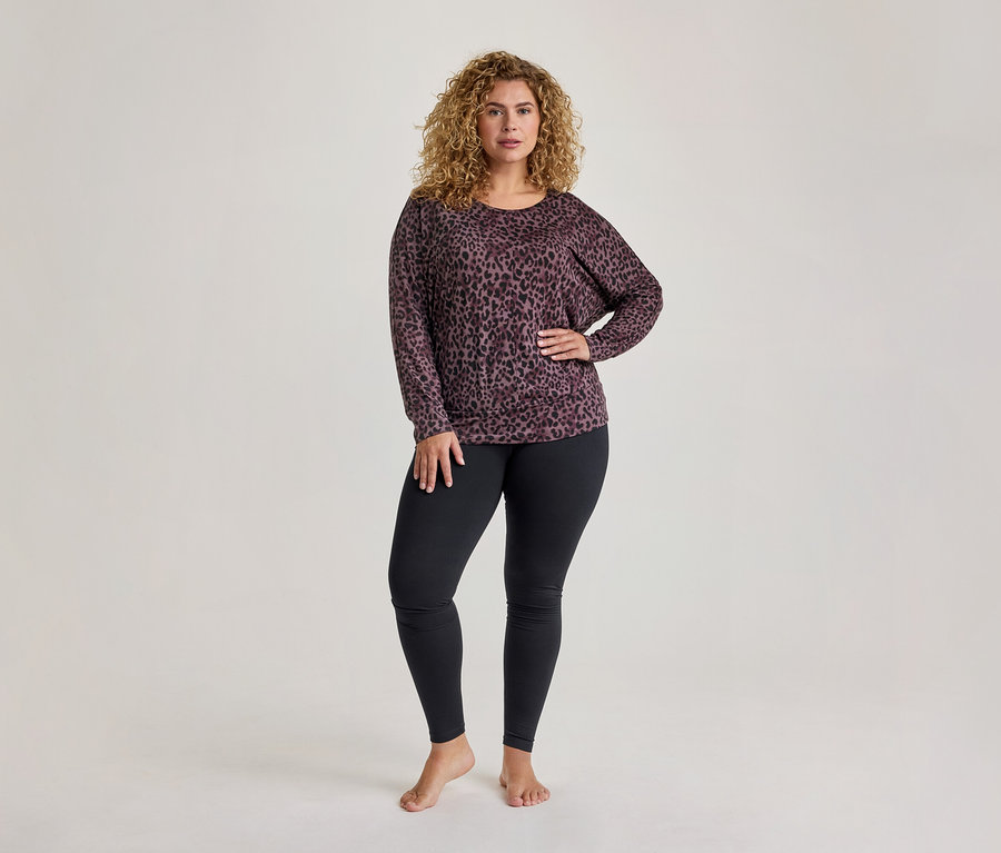 Eine Frau mit lockigem Haar steht barfuß, trägt ein langärmeliges Oberteil mit lila-schwarzem Leopardenmuster und schwarze Leggings, eine Hand in die Hüfte gestemmt.
