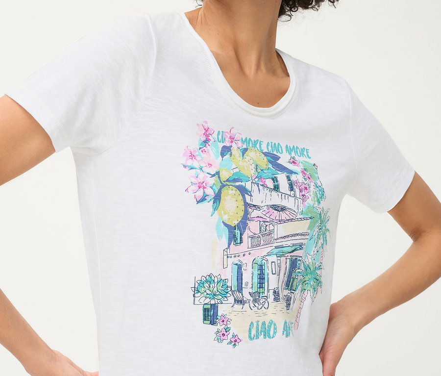 Person trägt ein weißes Olsen T-Shirt mit einem farbenfrohen Placement-Print, der ein mediterranes Gebäude, Zitronen, Blumen und Palmen zeigt.